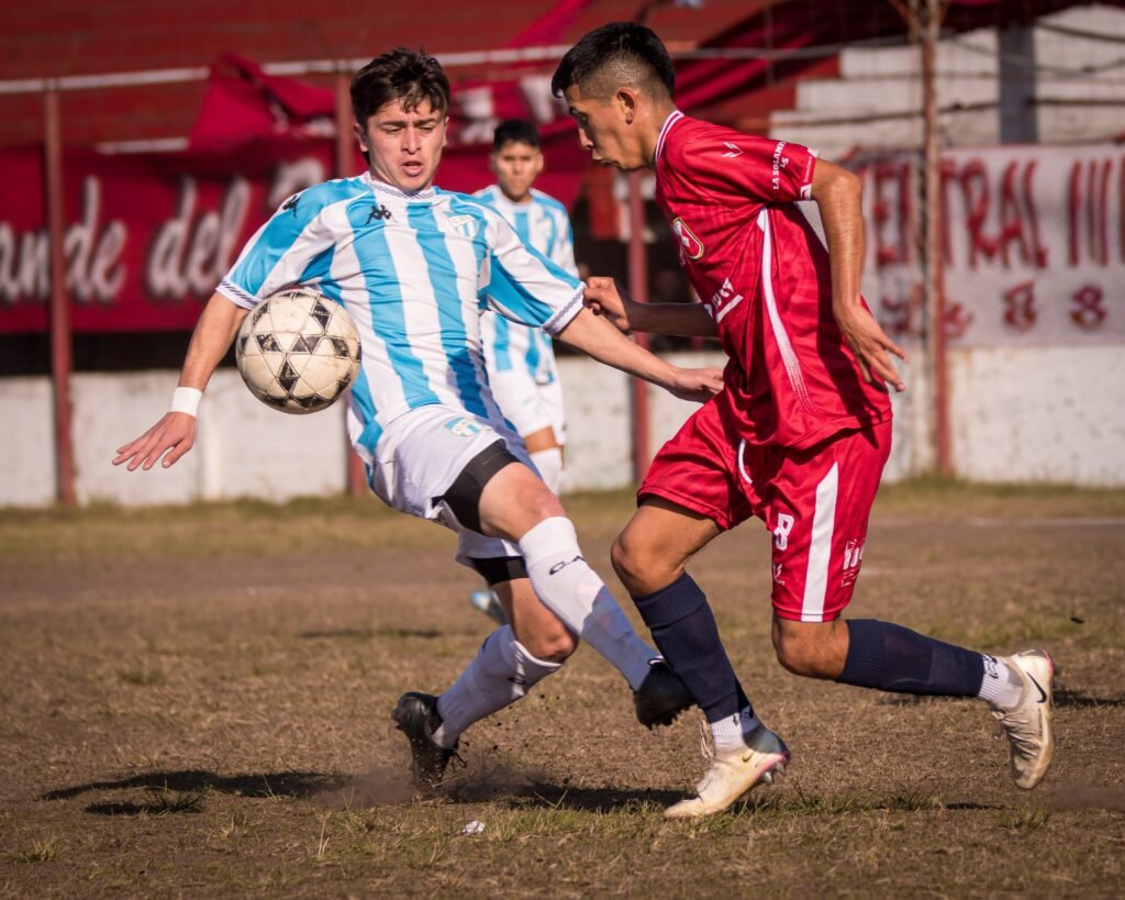 Jugador del equipo rival con camiseta a rayas celestes y blancas controla el balón, mientras un futbolista de Tucumán Central, vestido íntegramente de rojo, presiona de cerca en la marca sobre un campo de juego desgastado.