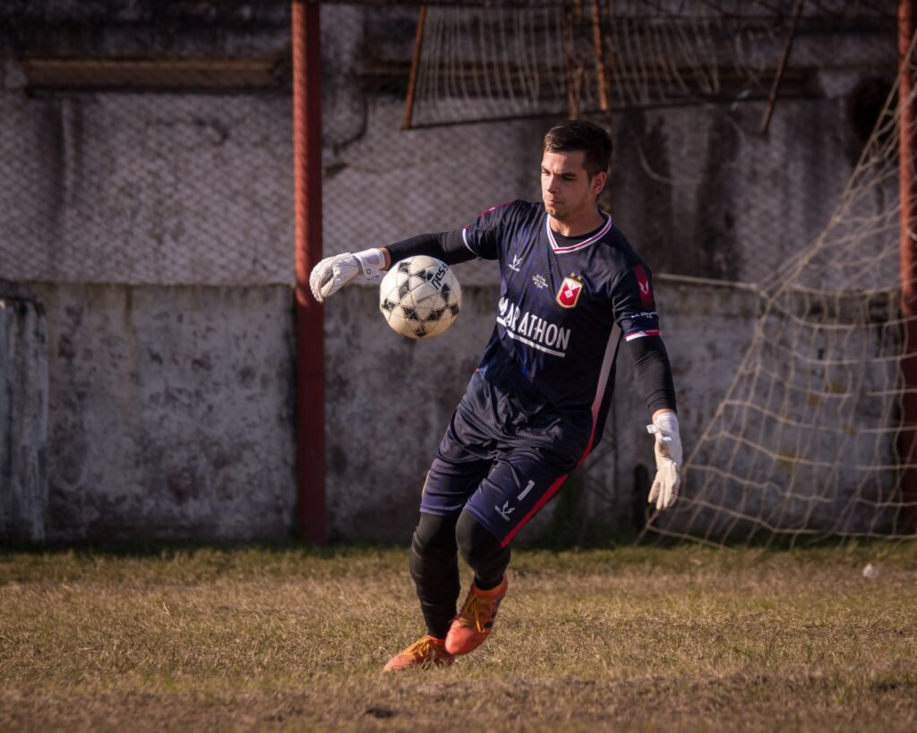 Arquero de Tucumán Central vistiendo conjunto azul oscuro con el logo de Marathon en el pecho, guantes blancos y botines naranjas, controlando la pelota con el pie derecho en la salida desde su área.