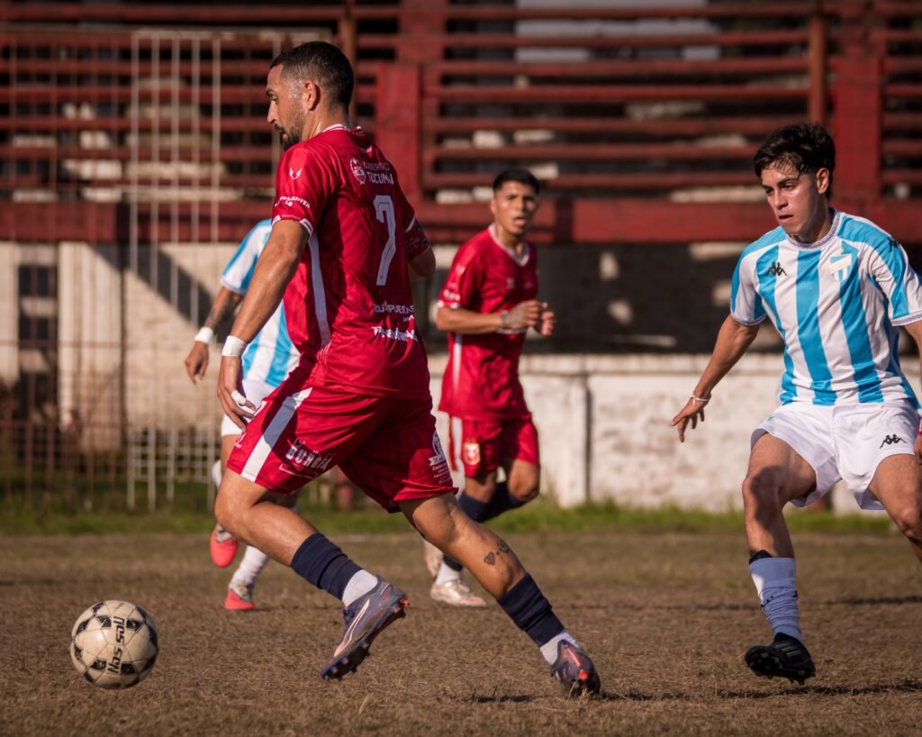 Jugador de Tucumán Central con indumentaria roja domina la pelota sobre el césped ante la marca de un rival con camiseta a bastones celestes y blancos.