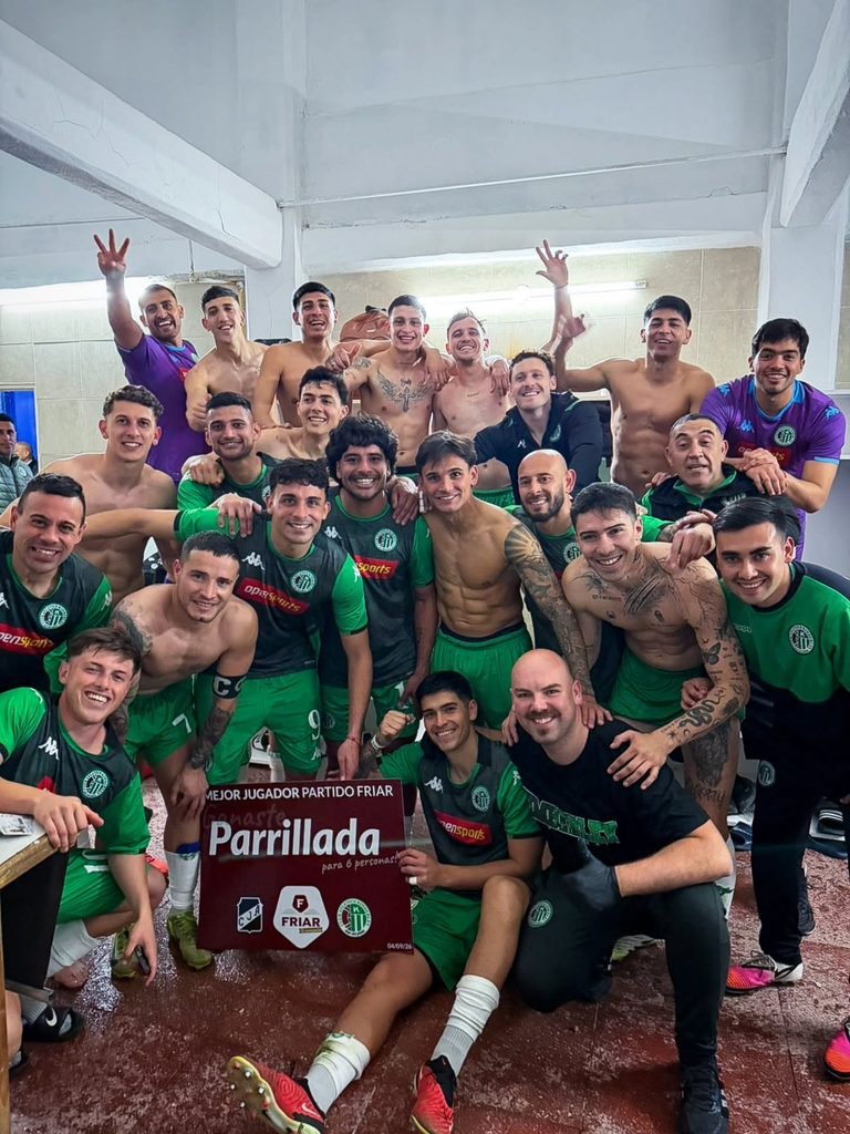 Plantel de Kimberley de Mar del Plata celebrando eufóricamente en el vestuario tras la goleada 3 a 0 sobre Juventud Antoniana. Los jugadores y el cuerpo técnico posan sonrientes abrazados, algunos con la indumentaria verde y negra del club, mientras un futbolista en primer plano sostiene un cartel rojo de premio al mejor jugador del partido.